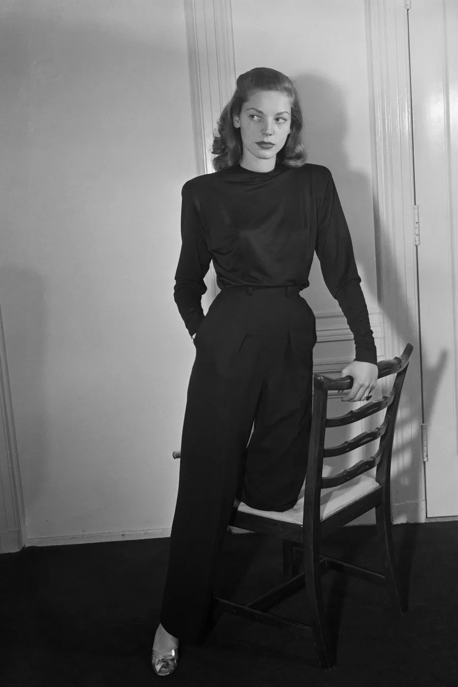 Black and white image of a model wearing a tailored black suit as she leans on a chair.