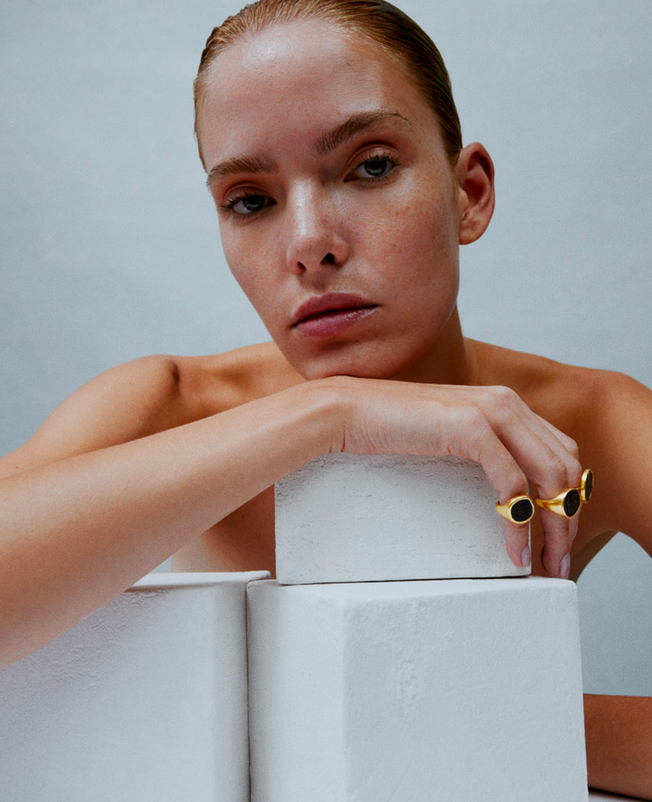 i seira model wearing three gold statement rings as she leans on a white block.