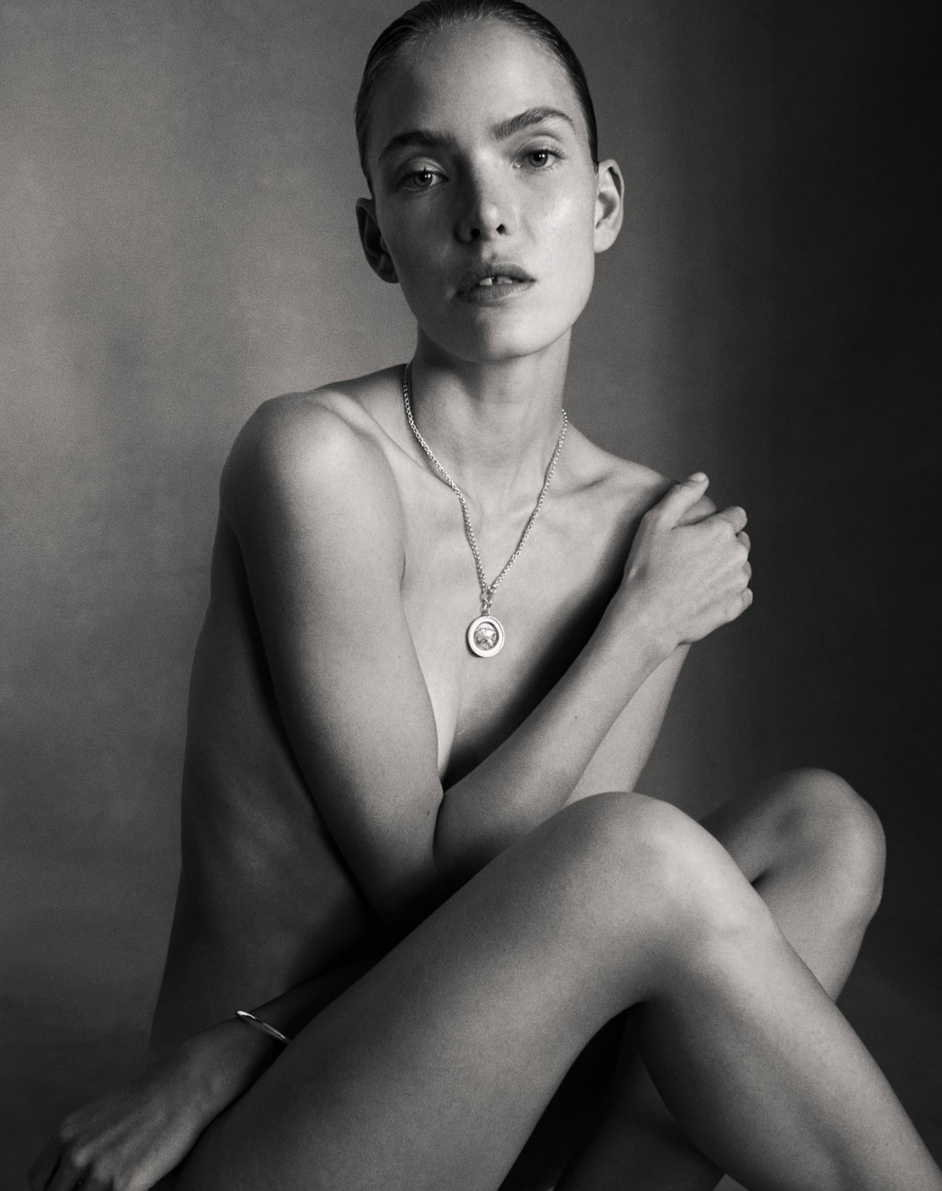 Black and white image of i seira model seated wearing only the reversible coin necklace with a gold bangle.